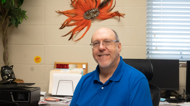 Professor Gil Cooper smiling in his office. He is wearing a blue shirt and he stands in front of a orange Venetian mask